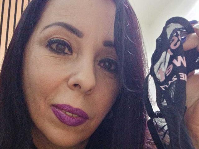 LauraSin, con cabello castaño oscuro, lápiz labial morado, sosteniendo una prenda interior de encaje negro con letras rosas, mirando a la cámara, sonriendo ligeramente, mostrando su nariz y labio superior..
