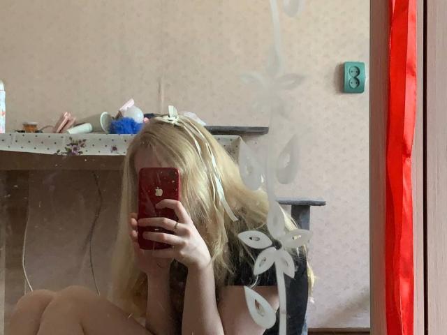 Hyacinth Bellin, loira, com flores no cabelo, vestindo uma blusa preta, tirando uma selfie no espelho com um celular vermelho, cobrindo o rosto, em um quarto bagunçado. O reflexo mostra uma moldura vermelha e manchas brancas..