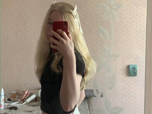 Hyacinth Bellin, con i capelli biondi, indossa una maglietta nera, sta scattando un selfie allo specchio con un telefono rosso, mostrando un piccolo accessorio per capelli bianco e un ripiano del bagno disordinato..