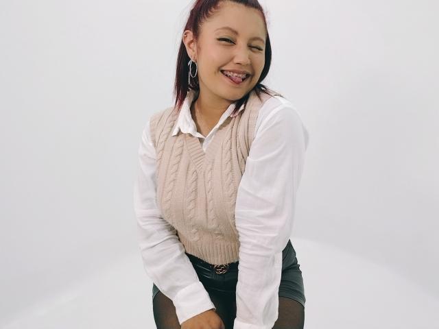 MarieHarper, smiling with red hair, wears a beige knitted vest over a white shirt, black skirt, and hoop earrings, standing with hands on knees, tongue playfully out.