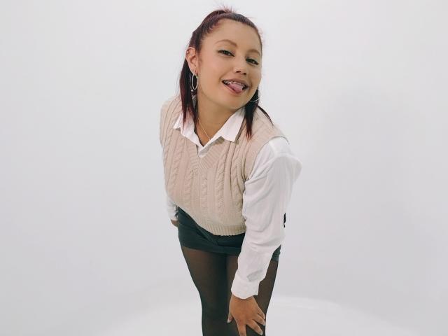 MarieHarper, with tongue out, wearing a beige knit vest, white shirt, black skirt, and black tights, leaning forward, smiling, and showing her small ass.