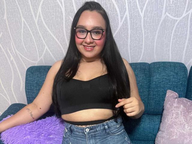 ValeryAndreas with long black hair, glasses, black sports bra, and blue jeans, smiling, showing a small tattoo on her left arm, sitting on a blue couch.