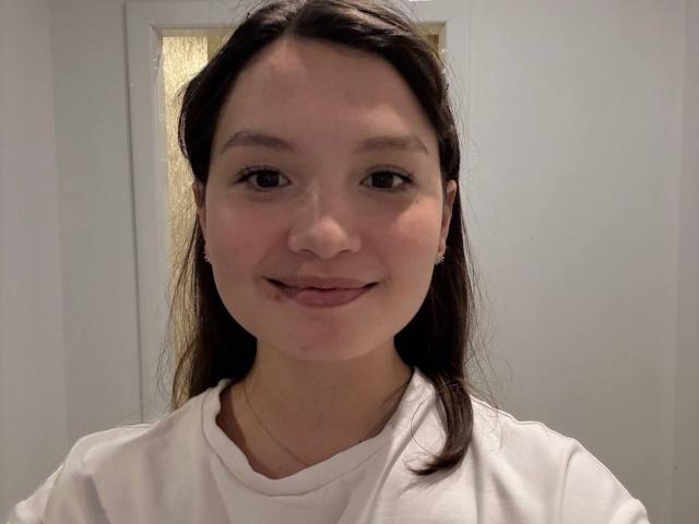 SuzanRhead, avec un sourire charmant, un petit nez, des cheveux bruns lisses, une chemise blanche et de discrètes boucles d'oreilles. Ses fesses ne sont pas visibles sur cette image..