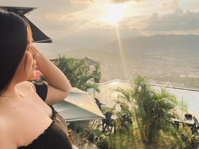Anny Bentleii, com cabelos longos e pretos, vestindo uma blusa preta, olhando para o sol, tem a mão na cabeça, e está em frente a um terraço com plantas e um guarda-chuva..