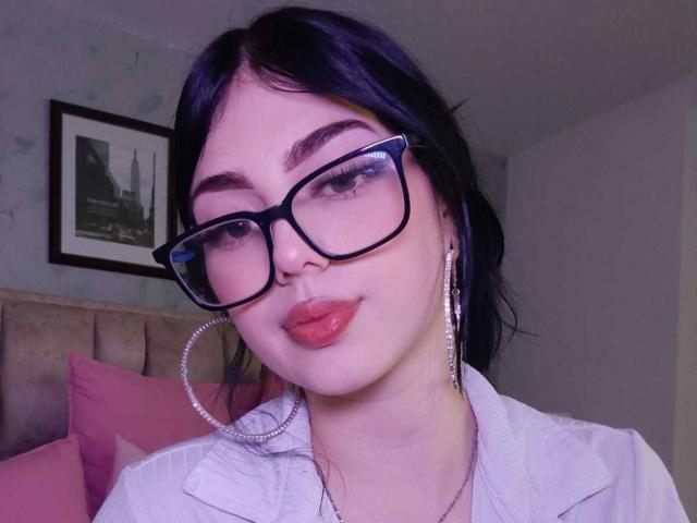 IsabellaaRossi with dark blue hair, wearing black glasses, sparkling hoop earrings, and pink lipstick, has a cute smile and white shirt. Her tits are small, and she's sexy.