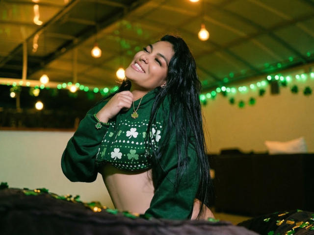 LannyShell with long black hair, smiling, wearing a green crop top with white clover patterns, exposing her tits, green string lights in the background, indoors, holding her shirt.