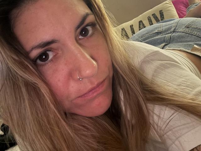 BellissimoFiore with long blonde hair, brown eyes, and a nose ring, lying down in a white shirt, showing a small part of her denim-covered ass in the background.