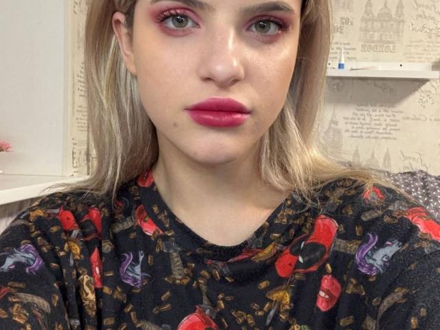 CoralieVonschriltz has pink eyeshadow, red lipstick, and a black shirt with red and purple cartoonish designs. Her blonde hair is down, and she's looking at the camera.