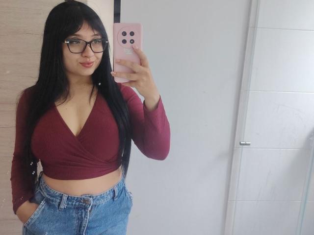 LunaBellaHotty with long black hair, glasses, red crop top showing her tits, blue jeans, holding a pink phone, taking a mirror selfie, smiling slightly.