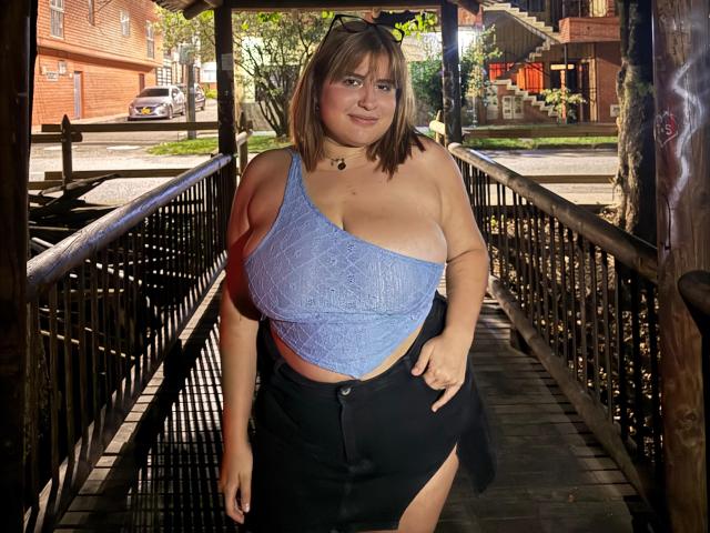 IvyOconner has big tits in a blue lace top, black skirt with a thigh slit, and a choker, standing in a wooden bridge, looking at the camera.