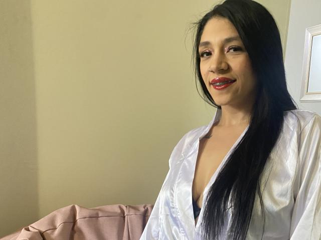 HoneyEvanss with long black hair, red lipstick, and a white silky robe, showing a bit of her black bra and small tits. She's smiling at the camera.