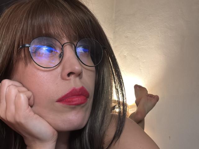 SeleneBright with glasses, red lipstick, and brown hair, resting her chin on her hand, showing her nose and subtle freckles, with her bare feet up in the background.