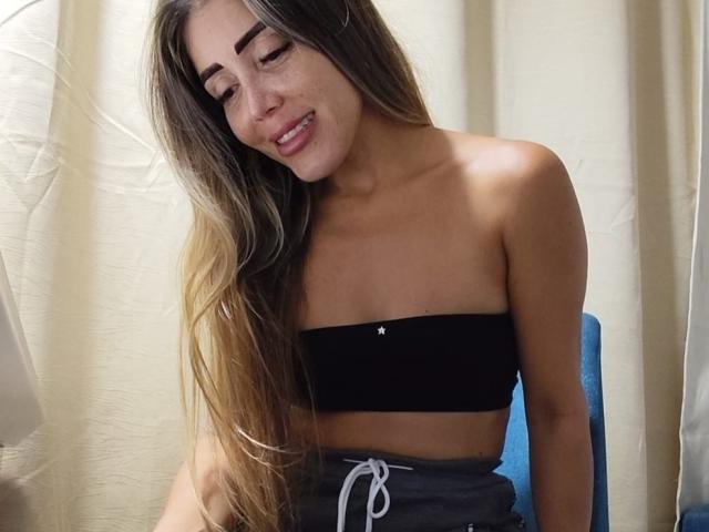 AndreaaHot with long, wavy brown hair, wearing a black strapless top, showing her small tits, and grey shorts, sitting and smiling, with a blue chair behind her.