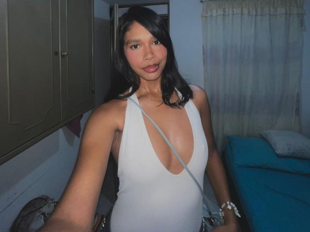 CamilaSwet, avec sa peau bronzée, ses cheveux noirs et son rouge à lèvres rouge, porte un haut blanc à encolure plongeante qui met en valeur sa poitrine généreuse, et se tient dans une pièce peu éclairée..