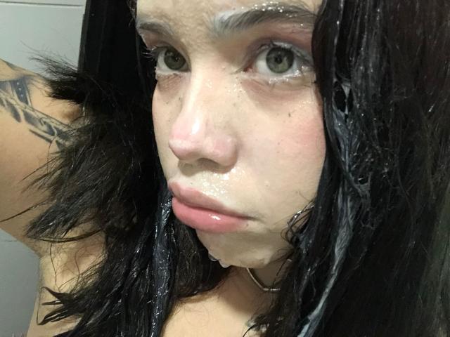 AryButty's wet face with black hair, white soapy residue on skin, gray eyes, pink lips, and a black tattoo on the left arm. She's wearing a black choker.