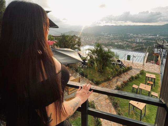AnnyBentleii with long black hair, small tits, tattooed arm, wearing a black top, standing by a balcony, looking at a sunlit, mountainous, city view, with green patio and tables.