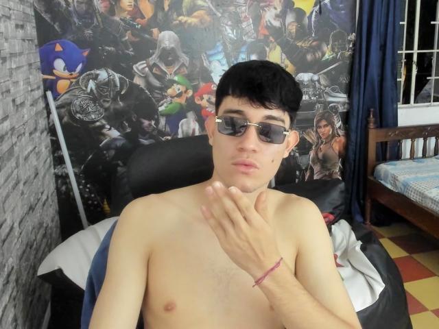 Shirtless JayOrange with black hair, glasses, and a pink bracelet, holding his chin, in a room with a Marvel poster. His hand is near his face.