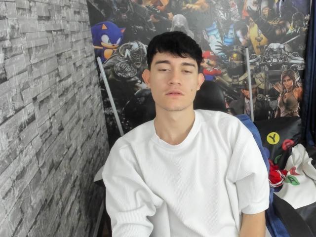 JayOrange, a young dude with short black hair, white shirt, and closed eyes, is sitting in a chair, looking bored with a small mole on his right cheek.