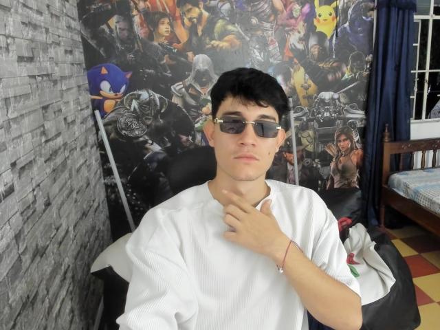 JayOrange with black hair, glasses, white tee, hand on neck, serious face, sitting in black chair, gray brick wall, colorful poster behind, red bracelet on right wrist.