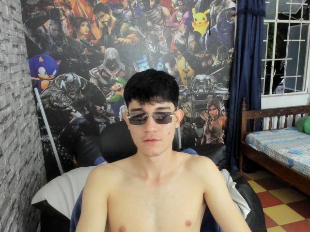 Shirtless JayOrange with black hair, glasses, and pale skin, sitting in a chair, wearing sunglasses, looking at camera, in a room with a colorful, comic-themed wallpaper.