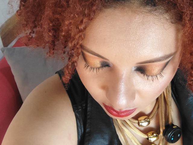 VioletaSwitch with red curly hair, golden eyeshadow, long eyelashes, red lipstick, black leather top, gold necklaces, and a black ring, looking down.