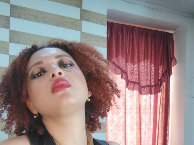 VioletaSwitch has curly brown hair, red lipstick, and dark eye makeup, looking up with a slightly open mouth. She's wearing gold earrings and a black top.