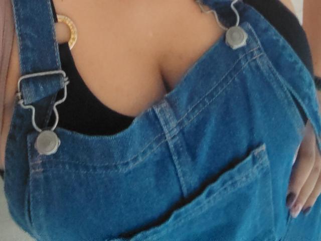VioletaSwitch's big tits are showing under a blue denim overall with metal rings, over a black top that's slightly visible. Her hand is on her right side.