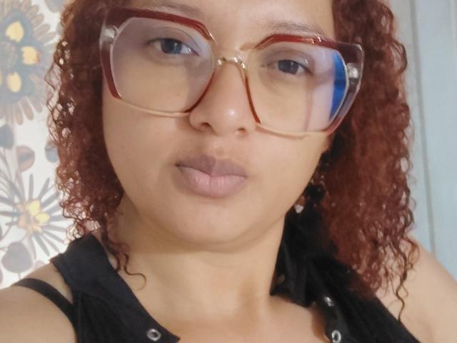 VioletaSwitch with curly red hair, wearing large red-framed glasses, black sleeveless top, and light brown lipstick, has a neutral expression, showing her nose and full lips.