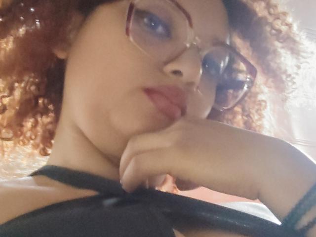 VioletaSwitch, con cabello rojo rizado, gafas y un sujetador negro de tirantes, muerde su dedo con una expresión sensual, mostrando sus labios carnosos y un atisbo de su sujetador negro..