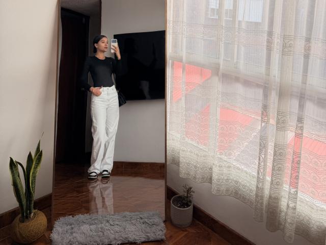ThaliaAres stands in front of a mirror, wearing a black top, white pants, black shoes, and a black cap, taking a selfie with her phone, showing her ass.
