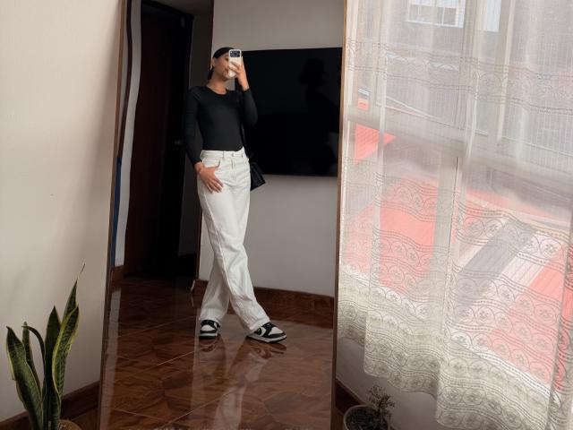 ThaliaAres, standing in a room, wearing a black long-sleeve shirt, white pants, black-and-white sneakers, holding a phone to her face, with a small plant in the corner.