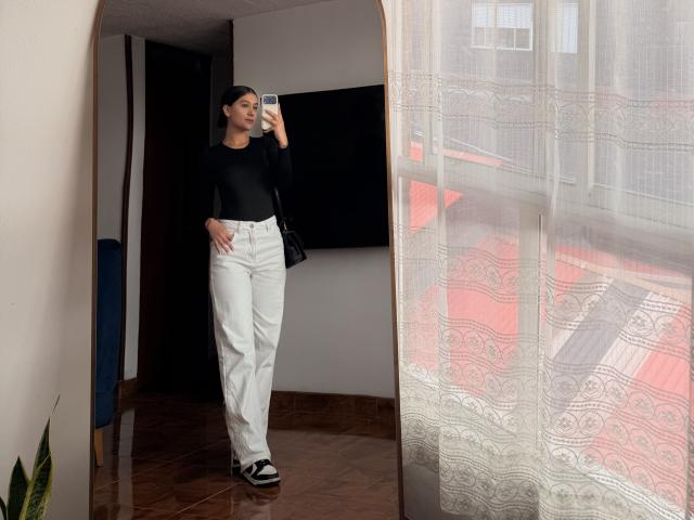ThaliaAres with short dark hair, black long-sleeve top, white high-waisted pants, black and white slip-on shoes, holding phone, standing in front of mirror, looking at reflection.
