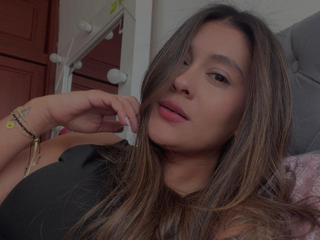 ThaliaAres has long brown hair, a cute face, pink lips, and is wearing a black top with a bracelet on her right hand, looking at the camera.