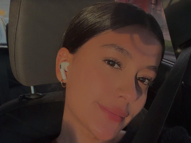 ThaliaAres with dark hair, white earbuds, and a slight smile, sitting in a car, wearing a black shirt, with a seatbelt across her chest.