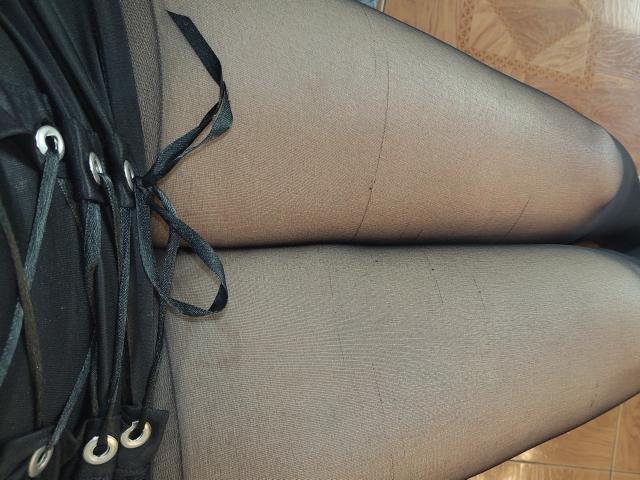 De zwarte, doorschijnende panty's van VioletaSwitch met een zichtbare naad en een zwarte rok met veter, die haar zwarte benen en zwarte linten laten zien, terwijl ze op een houten vloer zit..