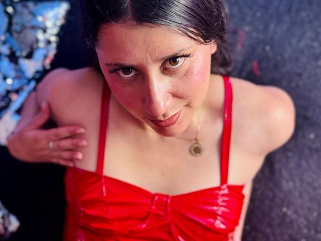 LunaDamasco with red shiny dress, looking up with brown eyes, wearing a gold necklace, her hand on her shoulder, sitting on a dark surface, short brown hair.