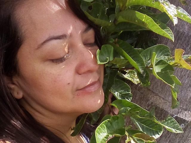 VonneLucia's face is wet with water droplets, eyes closed, leaning against green leaves with a slightly irritated expression. She has brown hair, light skin, and a small mole on her cheek.