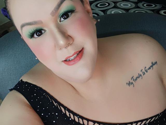 GiiannaMorrys with green eye makeup, nose ring, and tattoo on her shoulder, smiling at the camera, wearing a black, polka-dot top, showing her bare shoulders and chest.