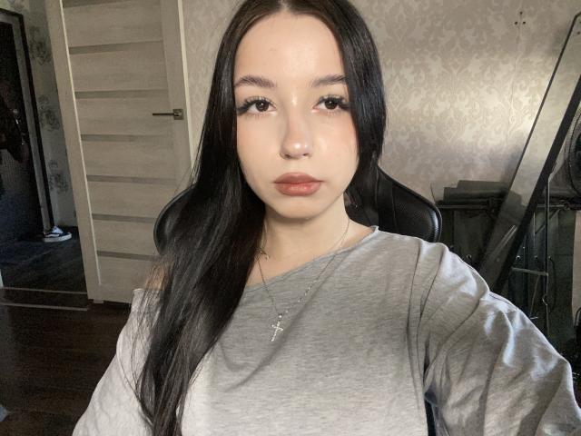 TaniaRaffa with long black hair, gray shirt, cross necklace, and subtle makeup, has a neutral expression, in a dimly lit room. Her ass is not visible.