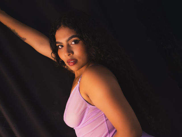 AshlyCorines with curly black hair, wearing a purple tank top, showing her tits, arm raised, tattoo visible, dark background, looking at camera, red lips, subtle makeup.