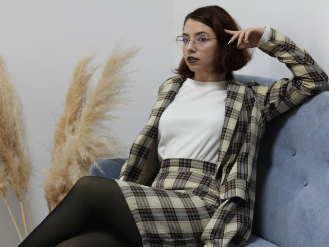 PolchaHodges mit Brille, kariertem Outfit, weißem Oberteil, schwarzen Strumpfhosen, sitzend auf einem blauen Sofa, Hand im Haar, leicht schmollende Lippen, braune Bob-Frisur, pralles Gesäß, Flanellrock, Pampasgras..