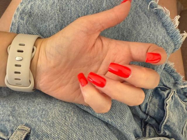 FellDivain's hand with shiny red nail polish, wearing a white smartwatch, resting on frayed blue denim. Her fingers are long and polished, showing off her red nails.