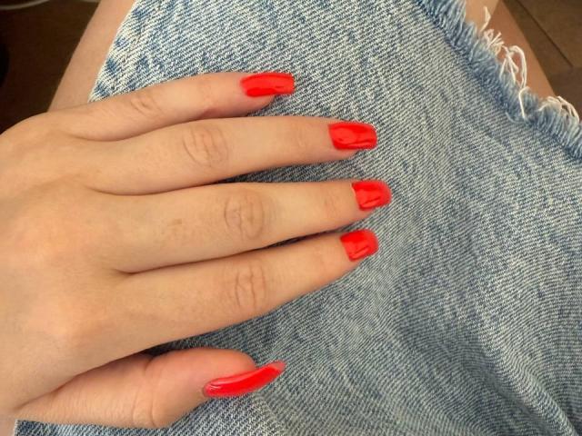A mão de FellDivain, com unhas vermelhas e brilhantes, repousava sobre o jeans azul desgastado. Seus dedos estavam ligeiramente afastados, exibindo suas unhas longas e brilhantes..