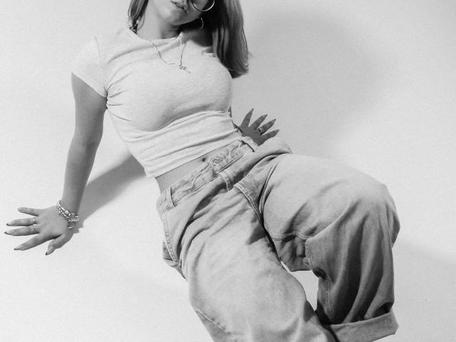 FellDivain, con un seno di media grandezza, indossa una maglietta grigia chiara aderente, jeans larghi, un braccialetto e occhiali, ed è appoggiata a un muro in una fotografia in bianco e nero..