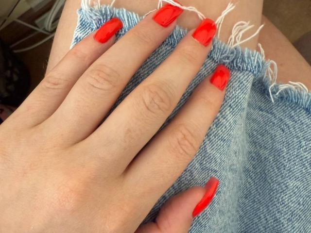 La main de FellDivain, avec ses ongles rouge vif, repose sur du denim usé. Ses doigts sont écartés, mettant en valeur ses ongles brillants et aux pointes carrées sur le tissu bleu texturé..