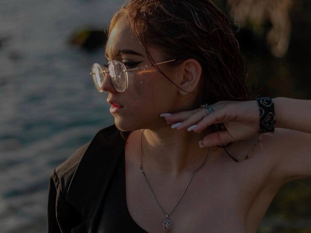 FellDivain, com cabelo castanho molhado, usando óculos de grau, uma camisa preta, um colar e uma pulseira preta, tocando o queixo à beira da água, mostrando seus seios em uma luz suave do final da tarde..