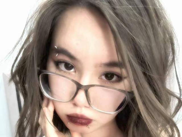 EmelineLattig with messy brown hair, wearing glasses, has dark lipstick, a nose piercing, and a lip piercing, looking at the camera with a slightly annoyed expression.