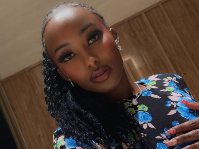 LustyMimi a la peau foncée, les cheveux noirs bouclés, et porte une robe fleurie avec des roses bleues. Elle regarde l'appareil photo avec des yeux séduisants à moitié fermés et porte un rouge à lèvres rouge foncé..