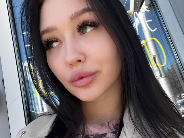 VannessaSchneidman with black hair, full lips, and long eyelashes, looking to the side, wearing a white coat, showing her cute face and pretty pink lipstick.