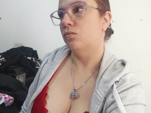 Amelia Wilson, avec des lunettes violettes, un sweat à capuche gris, un soutien-gorge en dentelle rouge, montrant ses grandes seins, un collier en argent avec un pendentif en métal, une expression légèrement boudeuse, des cheveux bruns attachés en chignon..